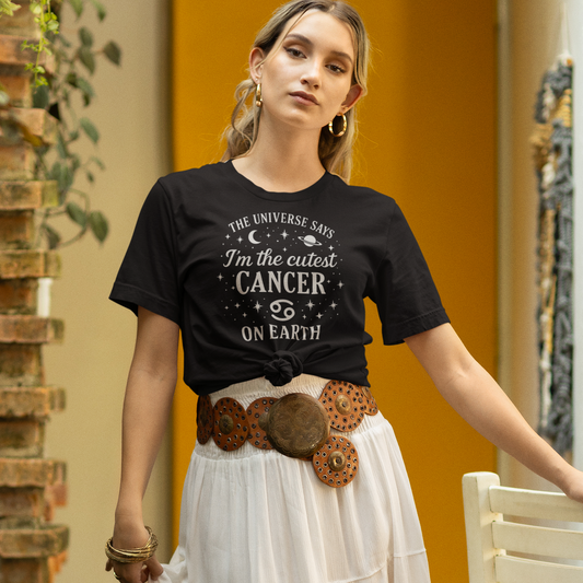 A woman wearing a black T-shirt with the white zodiac design that says “The Universe Says I’m the Cutest Cancer on Earth,” decorated with stars, a crescent moon and a planet. She is standing outdoors in a stylish boho outfit with a white skirt and wide belt, leaning on a chair.