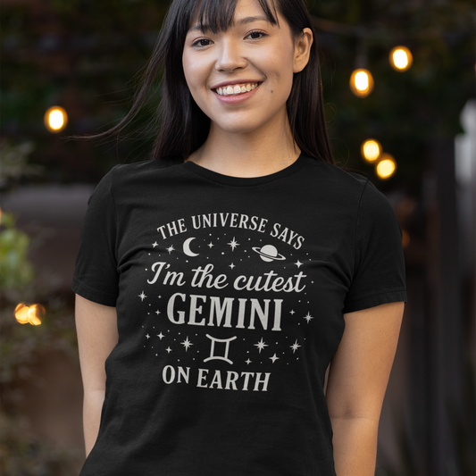 A smiling woman wearing a black T-shirt with a white Gemini zodiac design that says “The Universe Says I’m the Cutest Gemini on Earth,” decorated with stars, a crescent moon and a planet, standing outdoors with warm lights in the background.