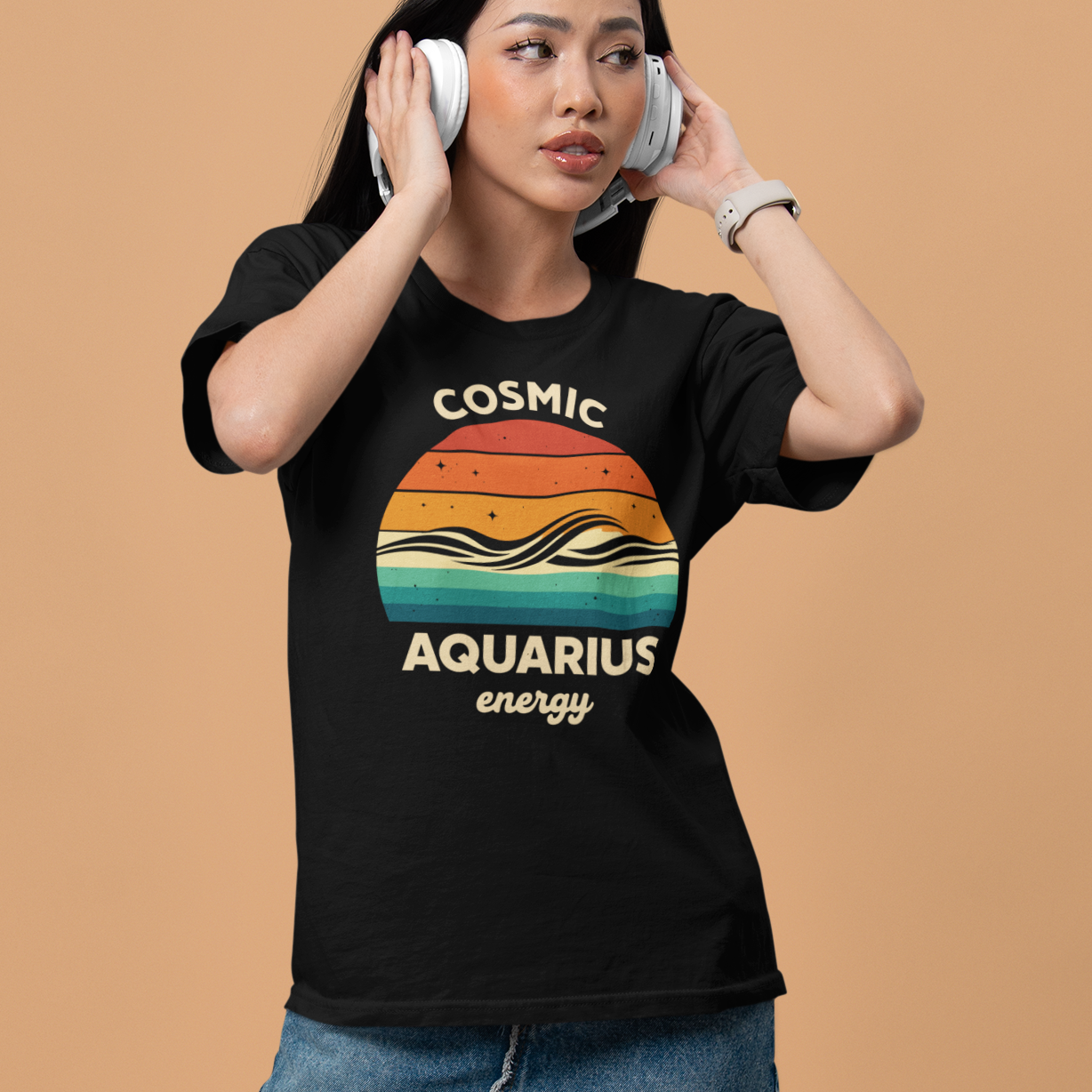 A young woman wearing a black T-shirt with a retro sunset-style graphic that reads “Cosmic Aquarius Energy,” featuring flowing wave lines and small stars. She is listening to headphones against a soft beige background.