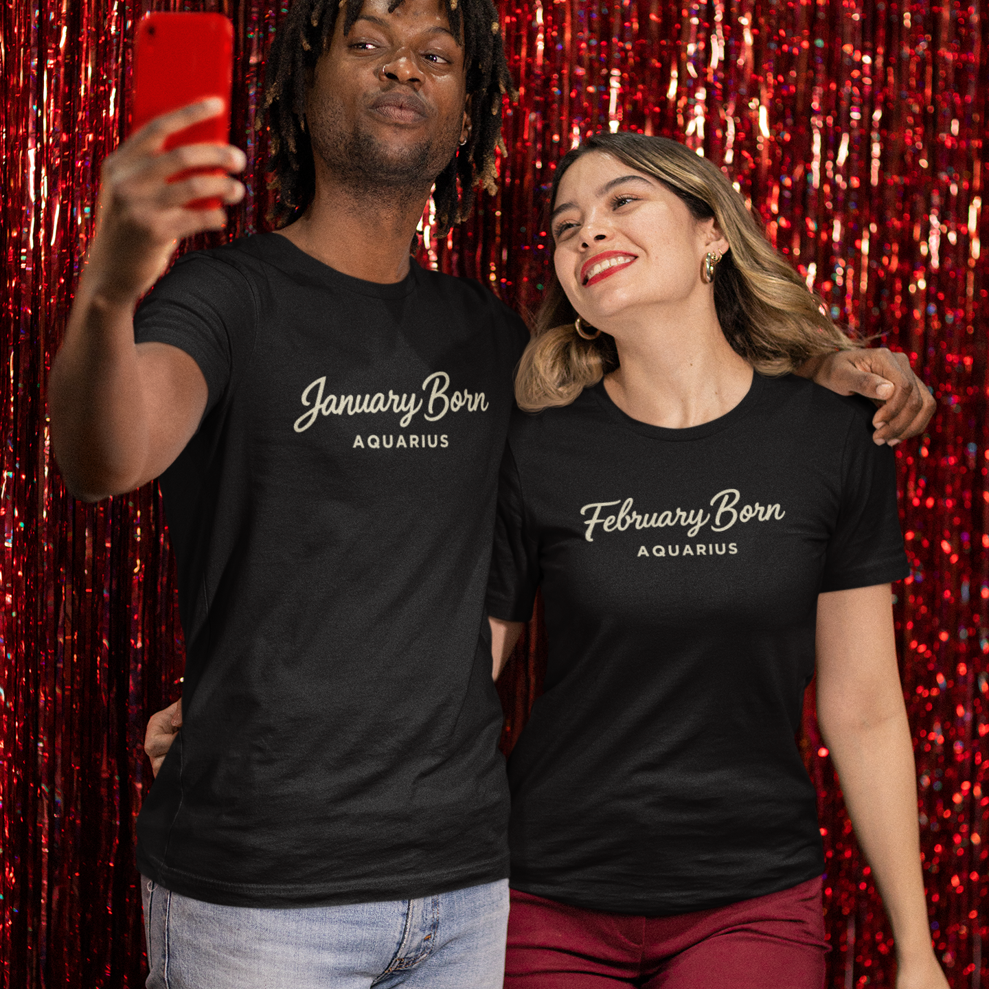 A man wearing a black January Born Aquarius T-shirt takes a selfie while smiling and standing next to a woman at a festive backdrop.