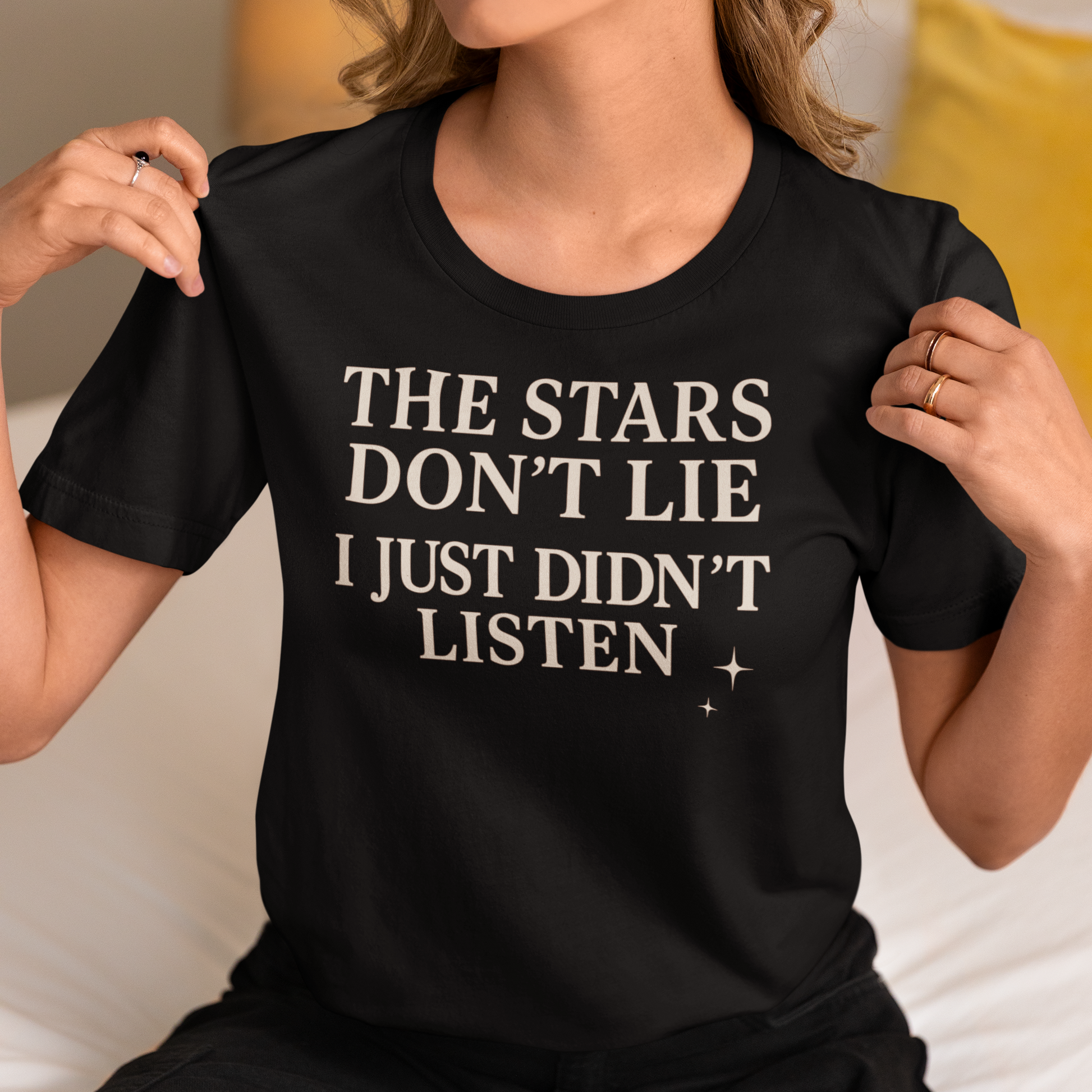 Woman wearing a black astrology T-shirt that says “The Stars Don’t Lie, I Just Didn’t Listen,” sitting on a bed and showing the design.