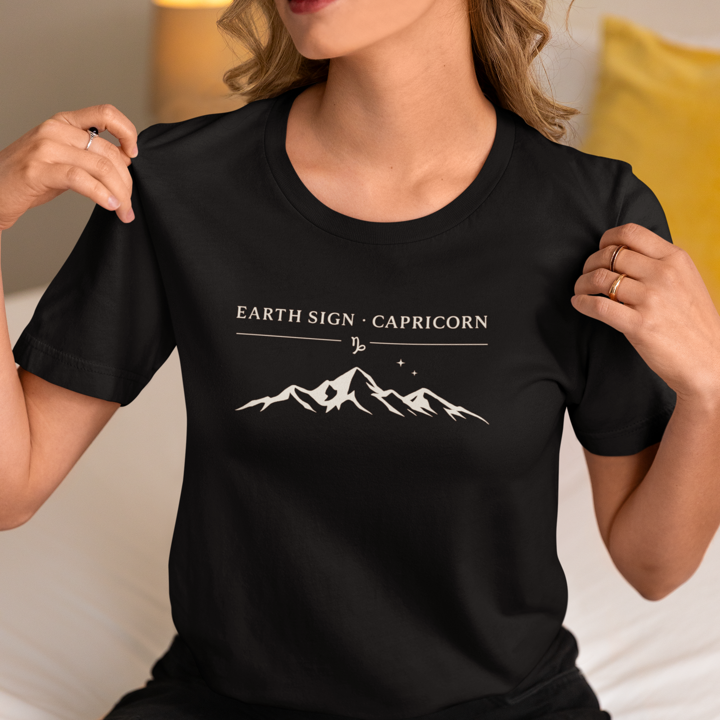Woman wearing a black T-shirt with the text “Earth Sign • Capricorn” and a minimalist white mountain graphic. Cozy indoor lifestyle photo.