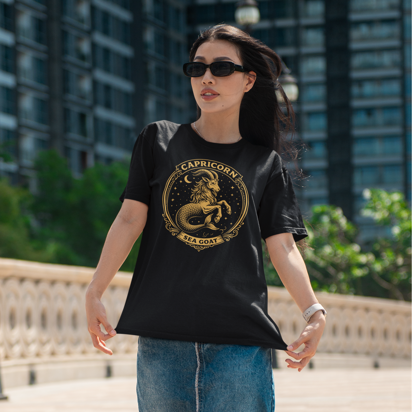 Woman wearing a black T-shirt with a vintage gold Capricorn Sea Goat zodiac design, standing outdoors in an urban city setting.