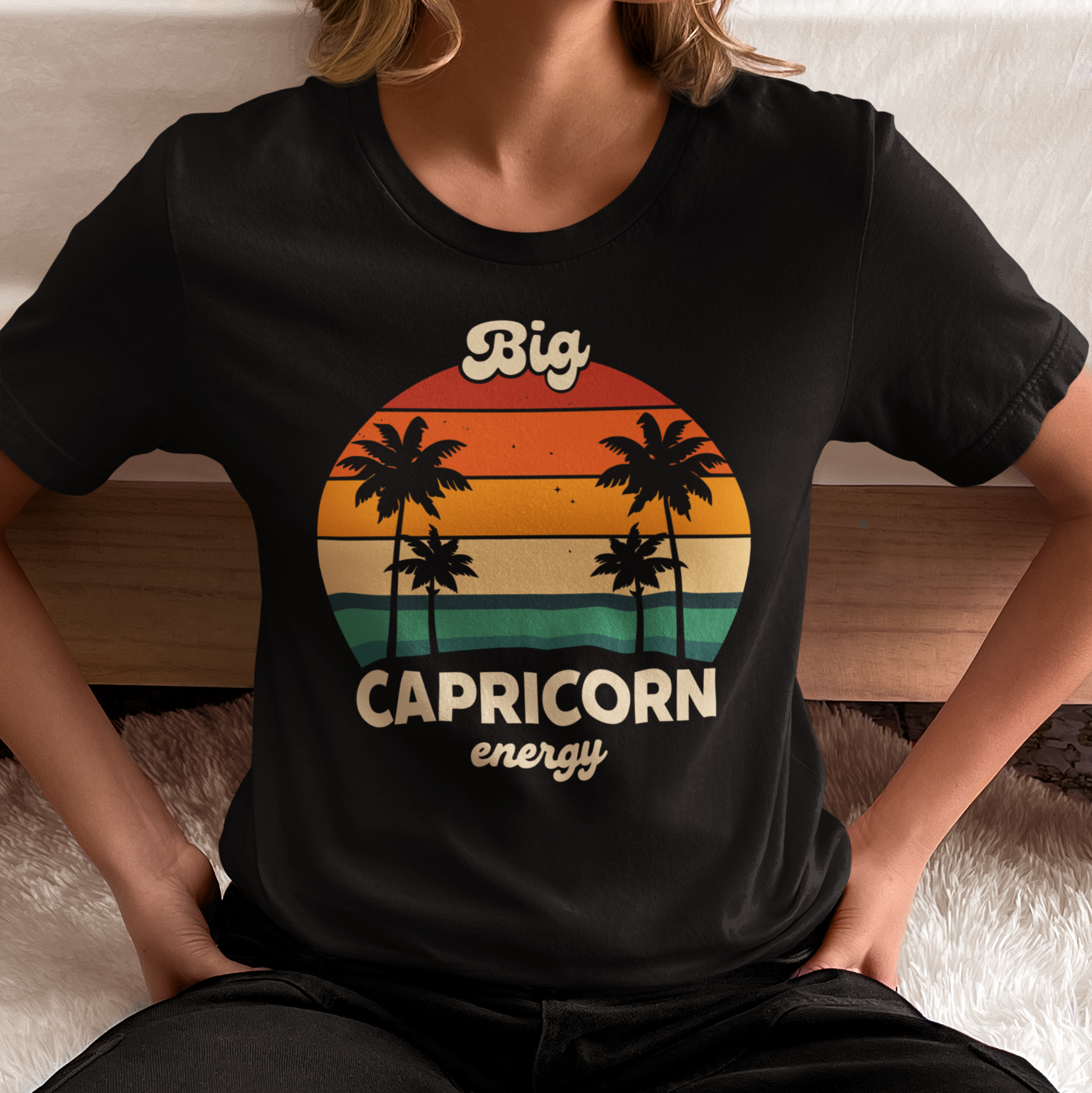 Woman wearing a black “Big Capricorn Energy” zodiac T-shirt with sunset and palm tree design in a relaxed indoor setting.