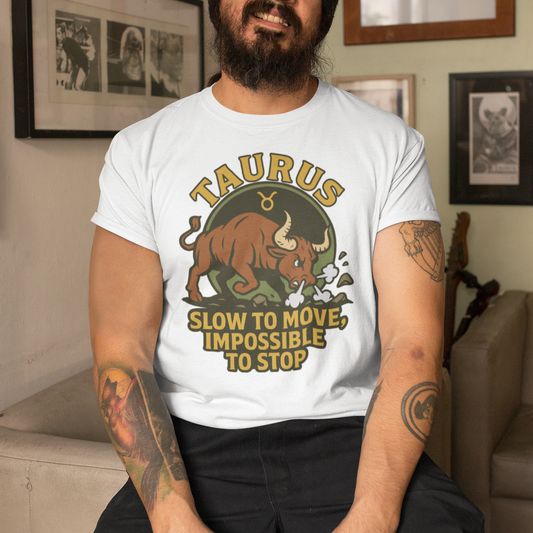 A smiling bearded man wearing a white Taurus T-shirt with a charging bull design and the text “Slow to Move, Impossible to Stop,” sitting indoors in a cozy room.