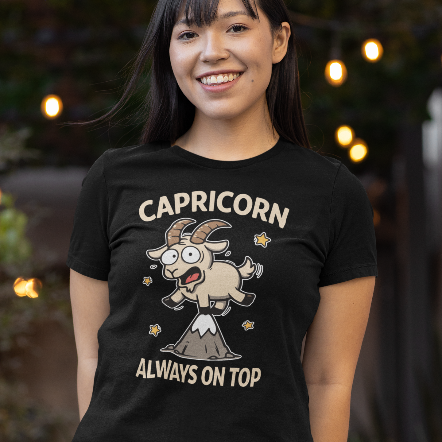 Model wearing a black Capricorn ‘Always On Top’ T-shirt featuring a playful cartoon goat standing on a mountain peak, photographed outdoors with soft lights in the background.