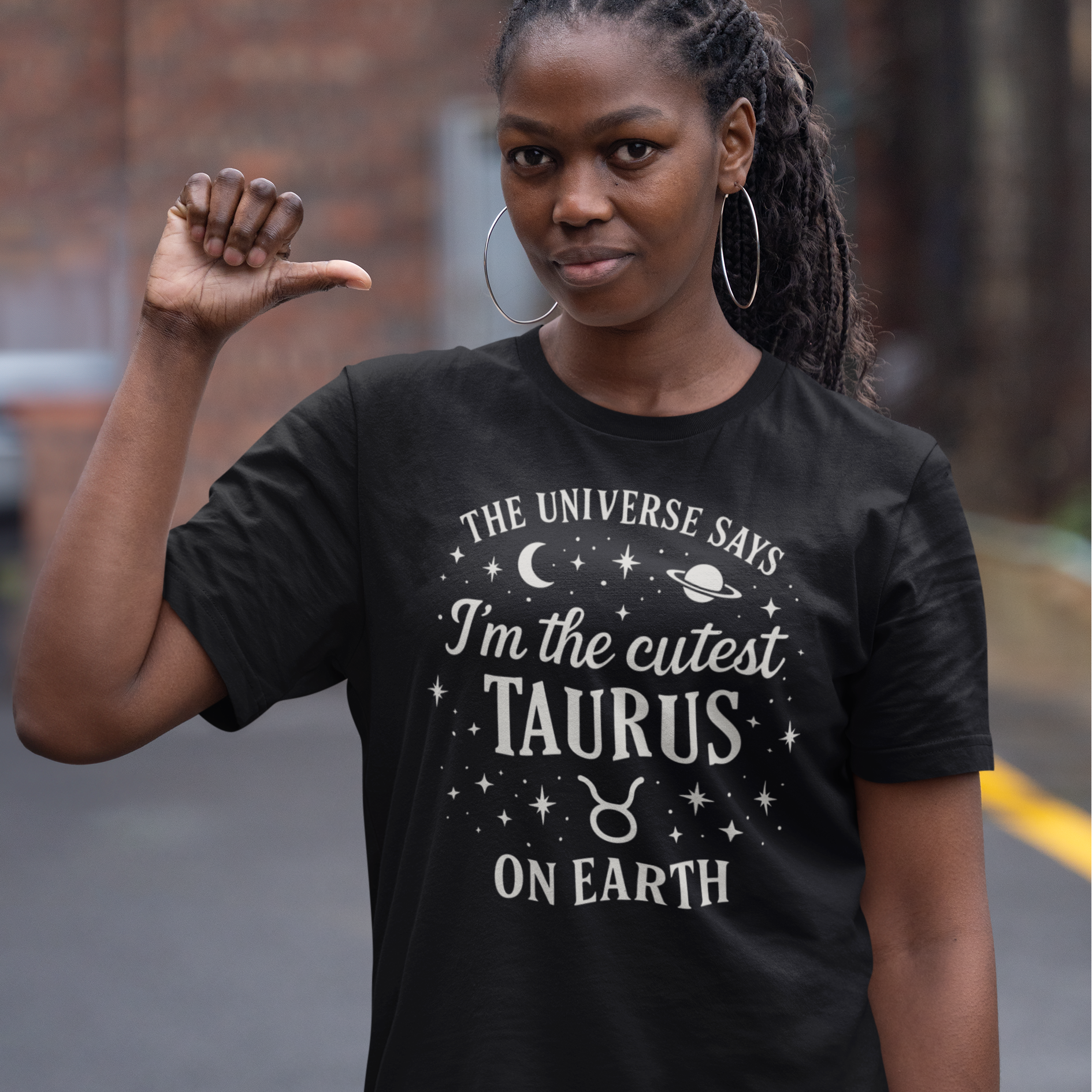 A woman wearing a black T-shirt with a white Taurus zodiac design that says “The Universe Says I’m the Cutest Taurus on Earth,” decorated with stars, a crescent moon and a planet. She is standing outdoors and pointing her thumb toward herself.