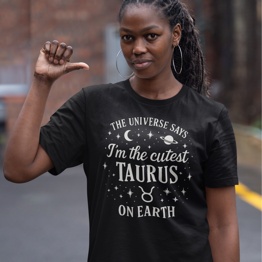 A woman wearing a black T-shirt with a white Taurus zodiac design that says “The Universe Says I’m the Cutest Taurus on Earth,” decorated with stars, a crescent moon and a planet. She is standing outdoors and pointing her thumb toward herself.