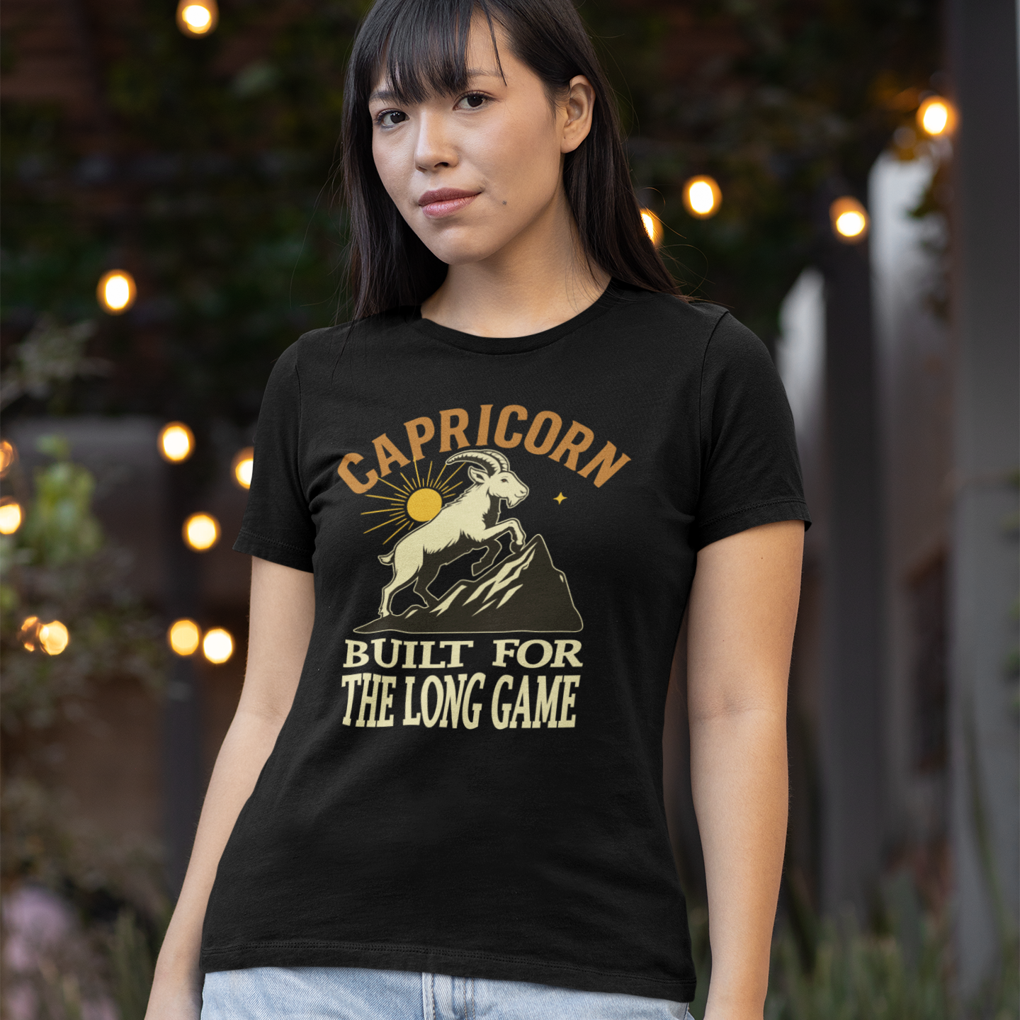 Model wearing a black Capricorn ‘Built for the Long Game’ T-shirt featuring a mountain goat illustration, photographed outdoors with soft lights in the background.