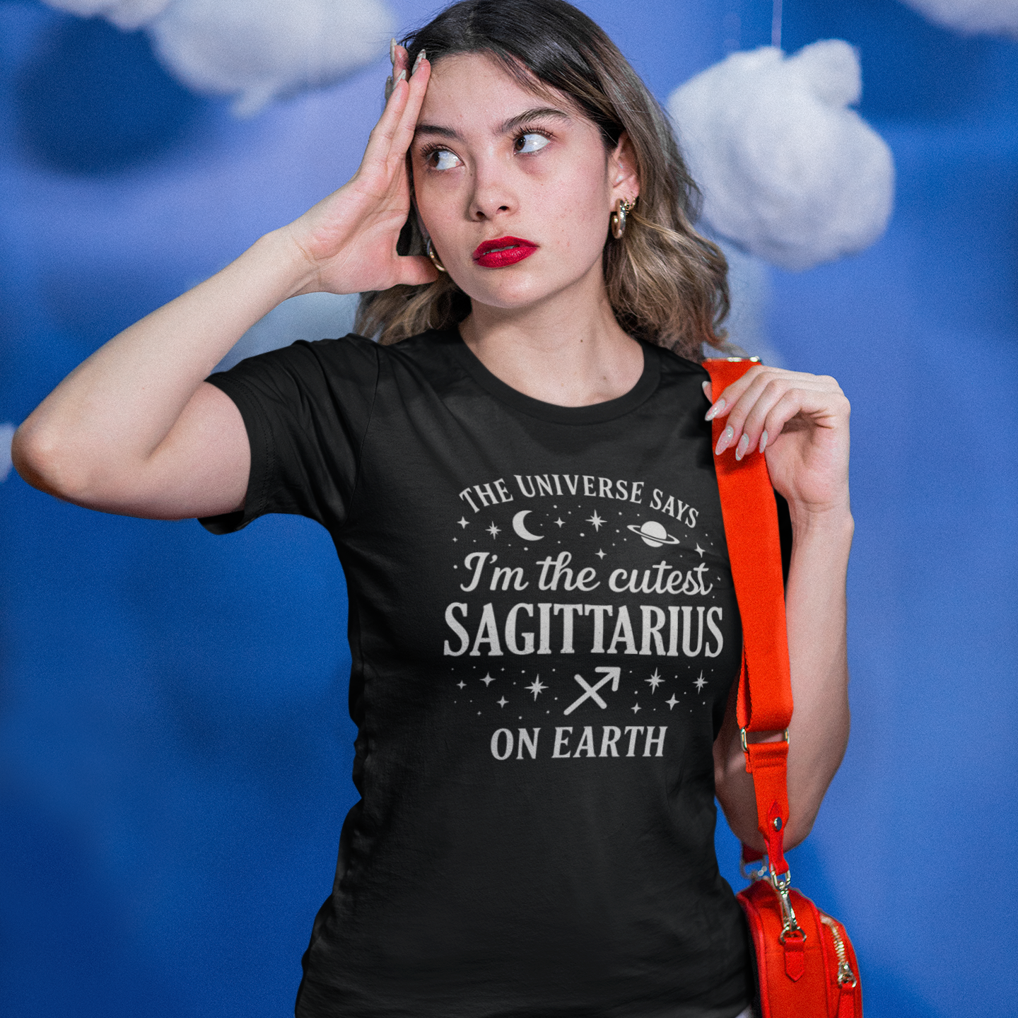 A young woman standing against a blue cloud-themed background wearing a black T-shirt with a white Sagittarius zodiac design that says “The Universe Says I’m the Cutest Sagittarius on Earth,” decorated with stars, a crescent moon, a planet and the Sagittarius zodiac symbol. She is holding an orange shoulder bag and touching her forehead as if thinking.